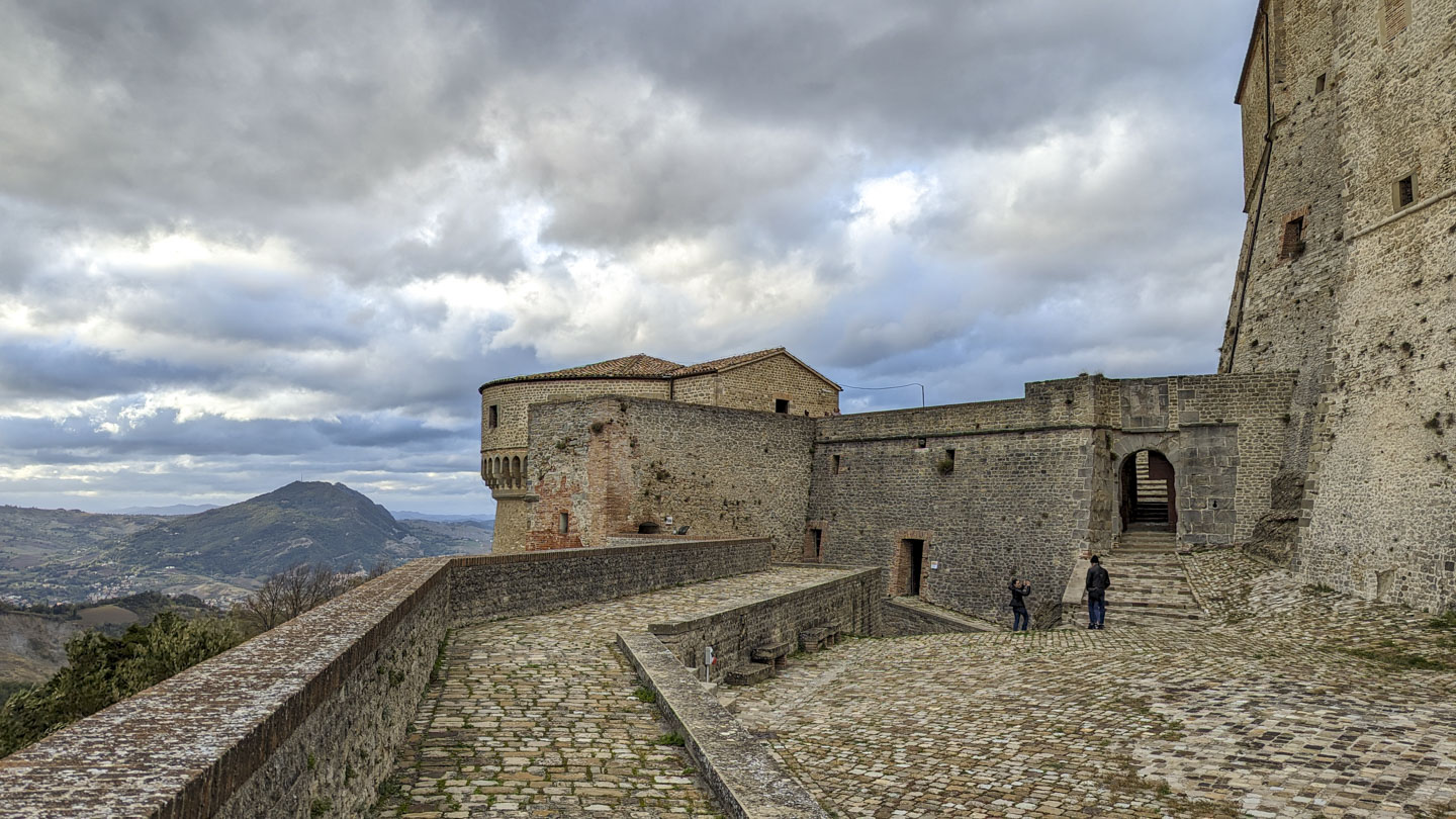 A mountain is on the left in the distance. A raised walkway leads away from you, and a lower plaza is on the right. Anne and Roberto are in the plaza, standing in front of a tall stone wall. The even taller walls of the fort are on the right.
