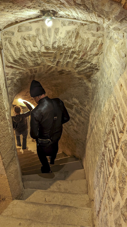 Anne and Roberto walk down a steep set of narrow stone steps with a relatively low ceiling.