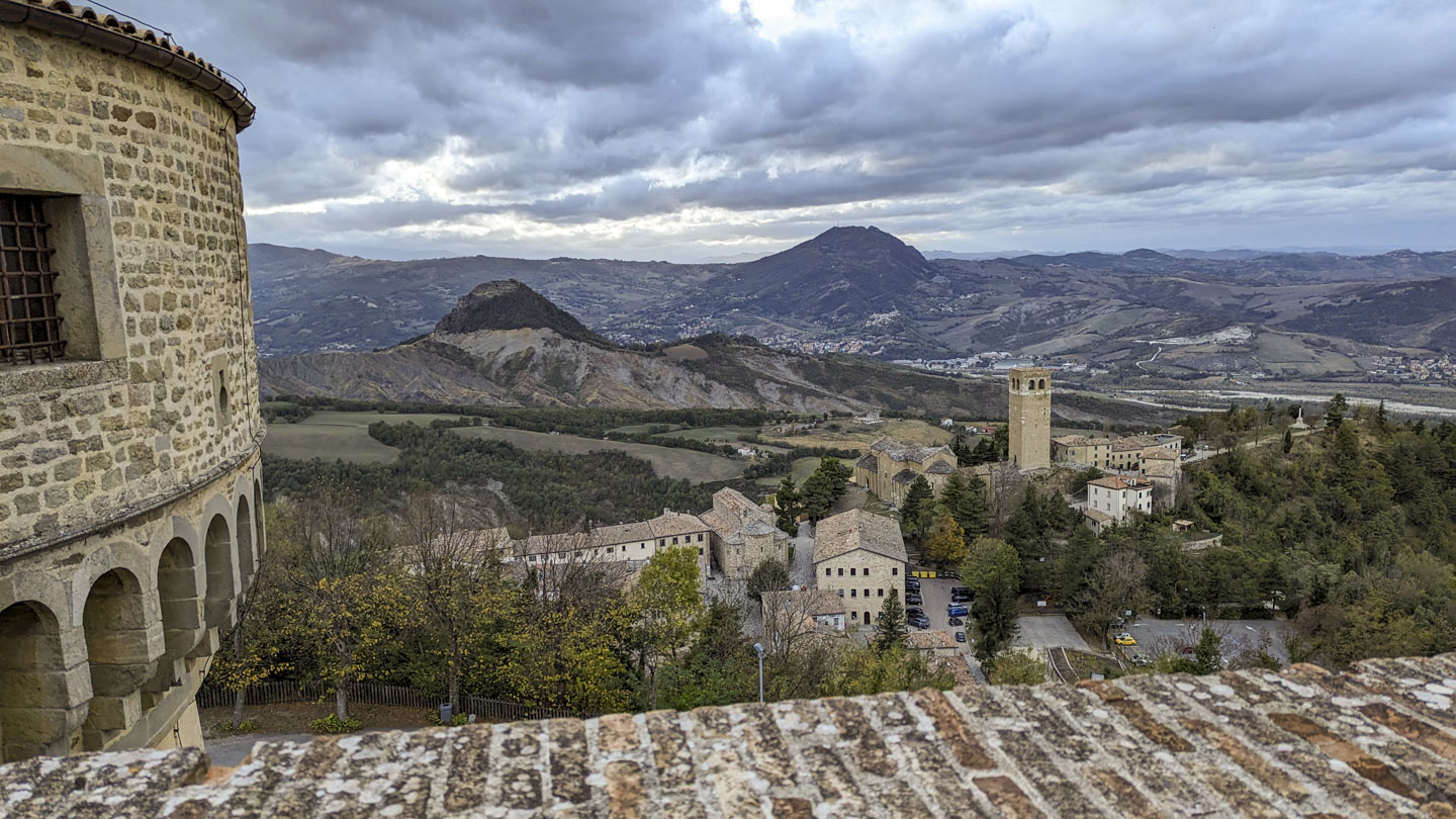 Photo taken over a brick wall, with a stone tower on the left. A village can be seen immediately below, with agricultural land and low mountains in the distance.