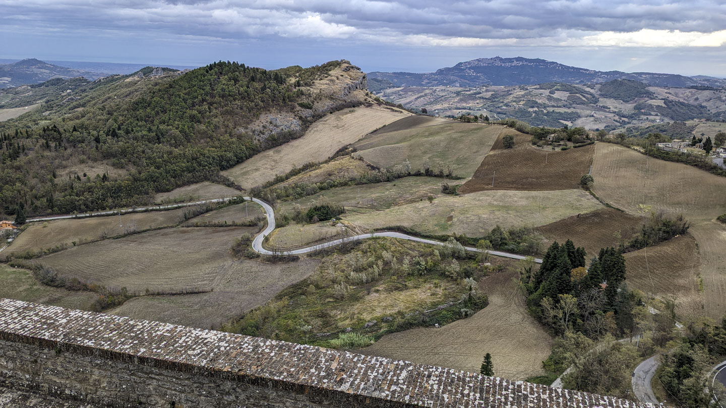 Taken over a brick topped stone wall, agricultural land on hills can be seen leading to San Marino.