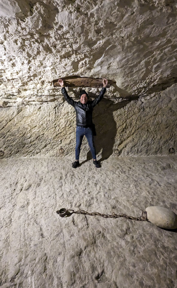 Roberto, wearing jeans and a black leather jacket, has his hands spread in the air against a wooden board that is embeded into the stone wall.