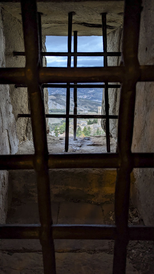 A window in a very thick wall. There are 3 sets of grates that are embedded into the stone. Through the window is a narrow view of the village and countryside far below.