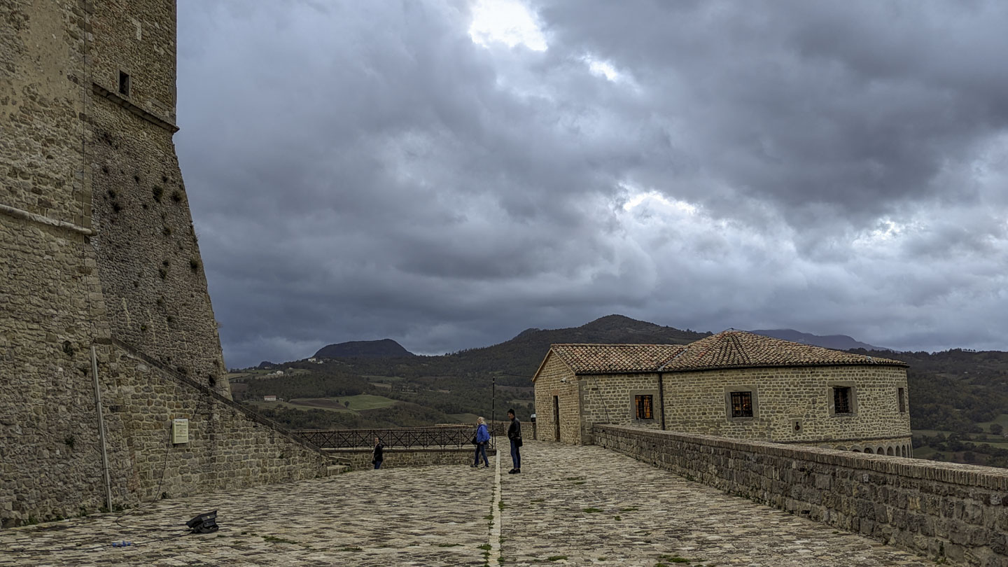 A tall stone wall is on the left, and a stone round building which is the top of a tower is on the right. Three people are in the center, and look small against the buildings.