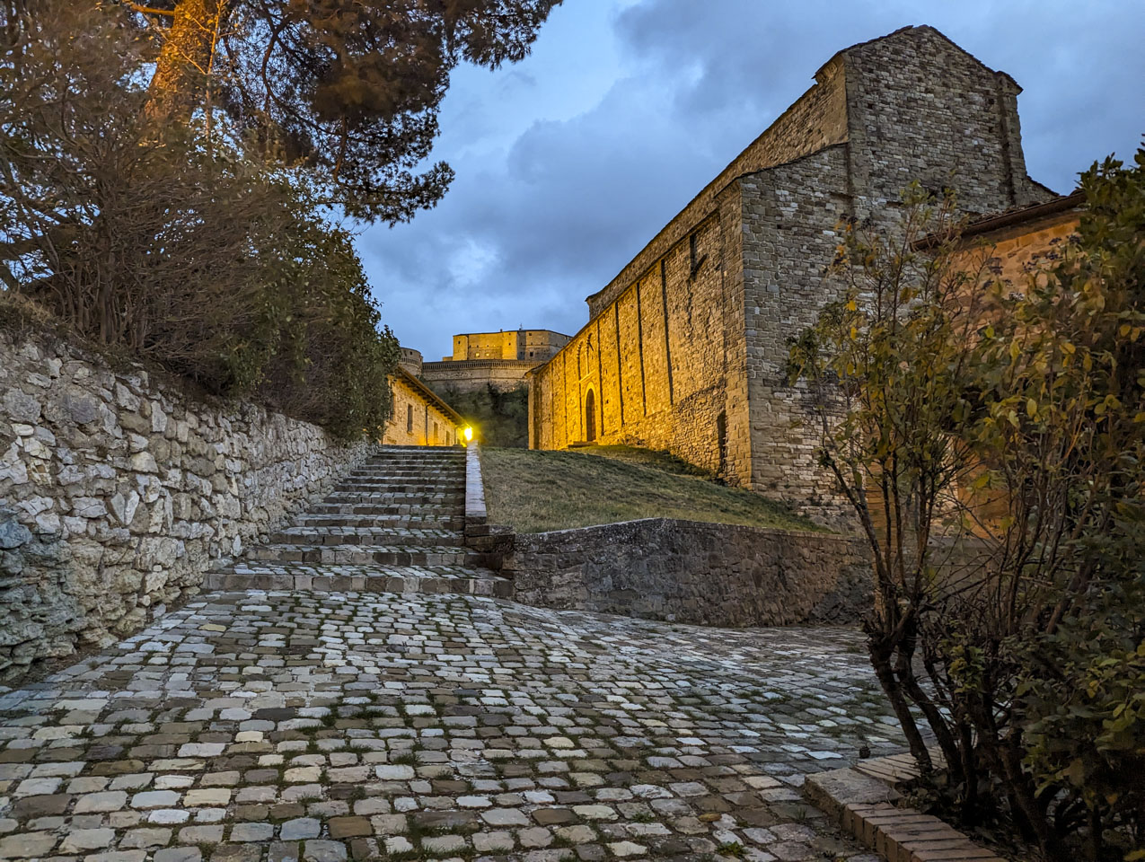 Looking across a coblestone walkway up stairs that use the same stone. In the distance at a high elevation is the fortress. Closer we see the stone walls of an old church.