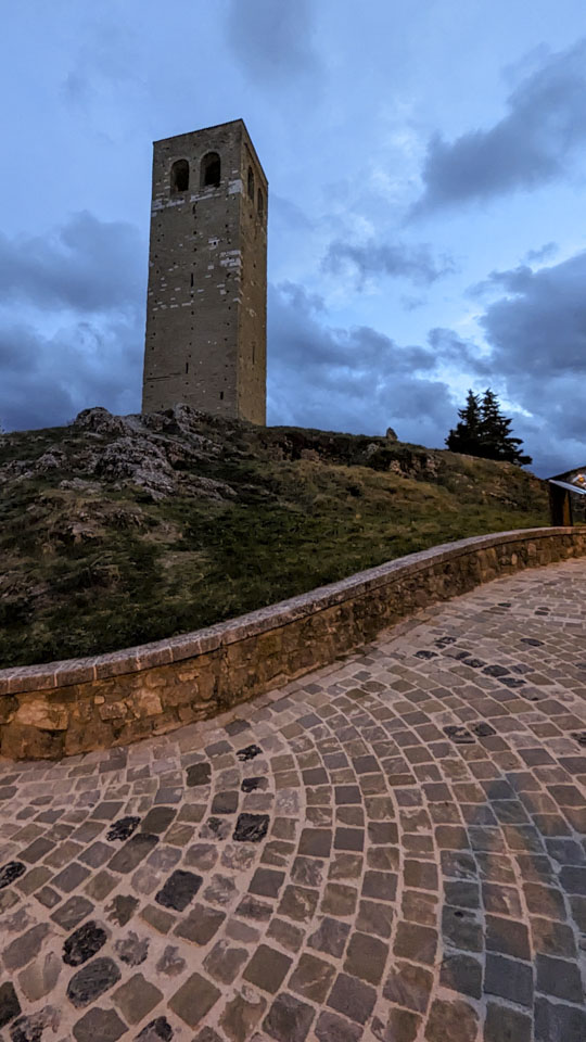A stone tower is on the left of this photo, with a partially cloudy dusck sky above it. There is a slight glow from streetlights on the curved stone walkway in the foreground.