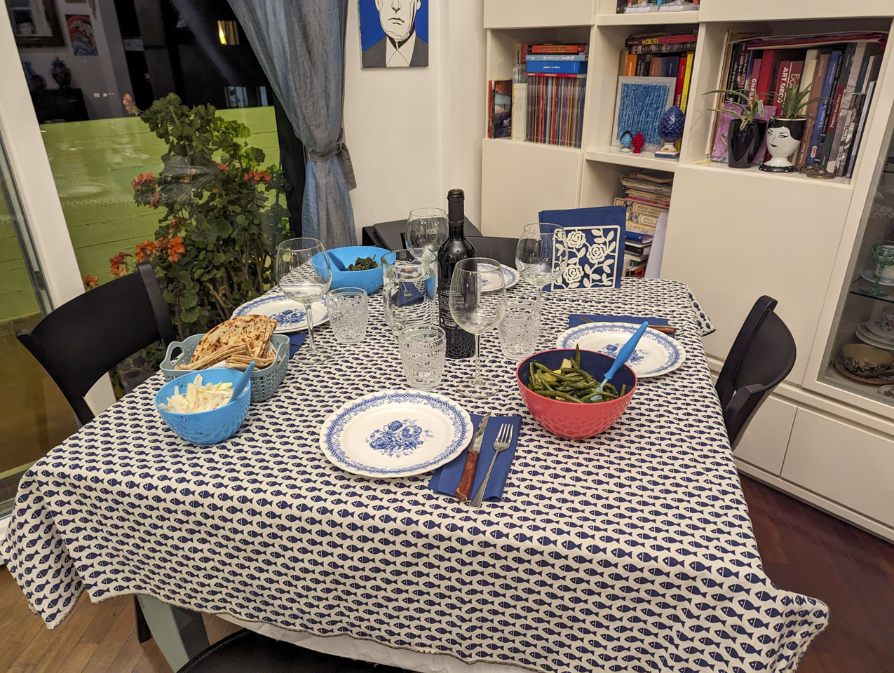 A table with a fish tablecloth. There are 4 empty plates, 4 empty wine glasses, 4 empty water glasses, and a pitcher of water. An open bottle of wine is in the center, and there are a few containers with food also on the table. The wall on the right has a bookshelf, while the wall on the left is a glass sliding door.