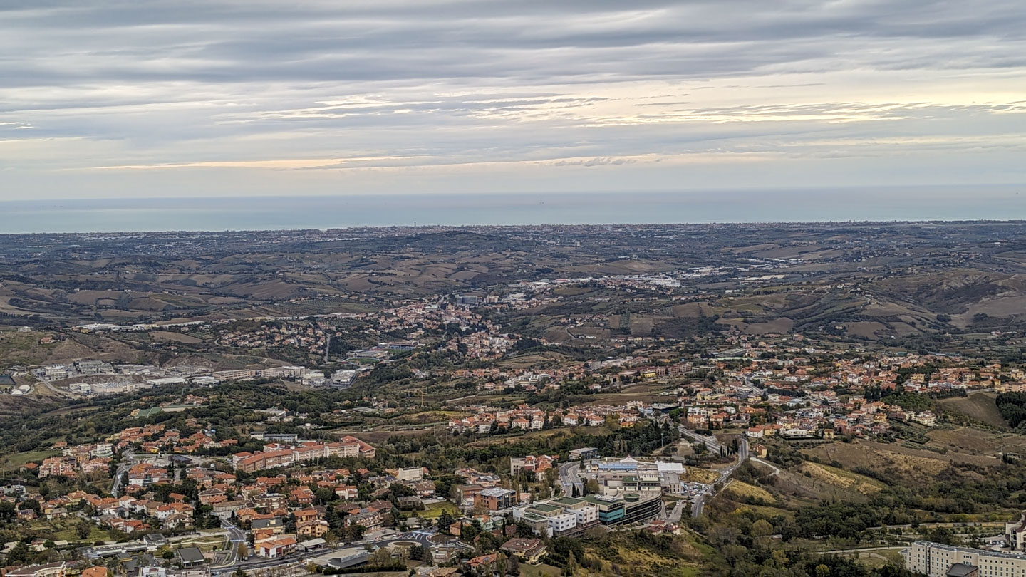 Photo taken from a high position showing an expanse of land that is relatively flat. There are some agricultural fields and many settlements. In the distance the Adriatic Sea can be hazily seen.