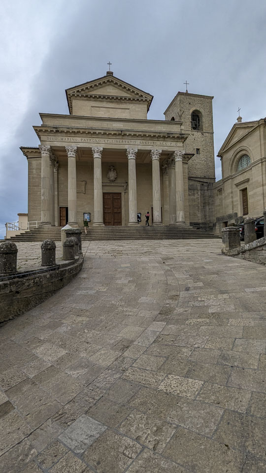 A wide stone path leads up to a plaza. On the far side of the plaza are stairs that lead up to a large building with 6 pillars in front. There is a cross at the top of the building, and on the tops of two adjacent buildings.