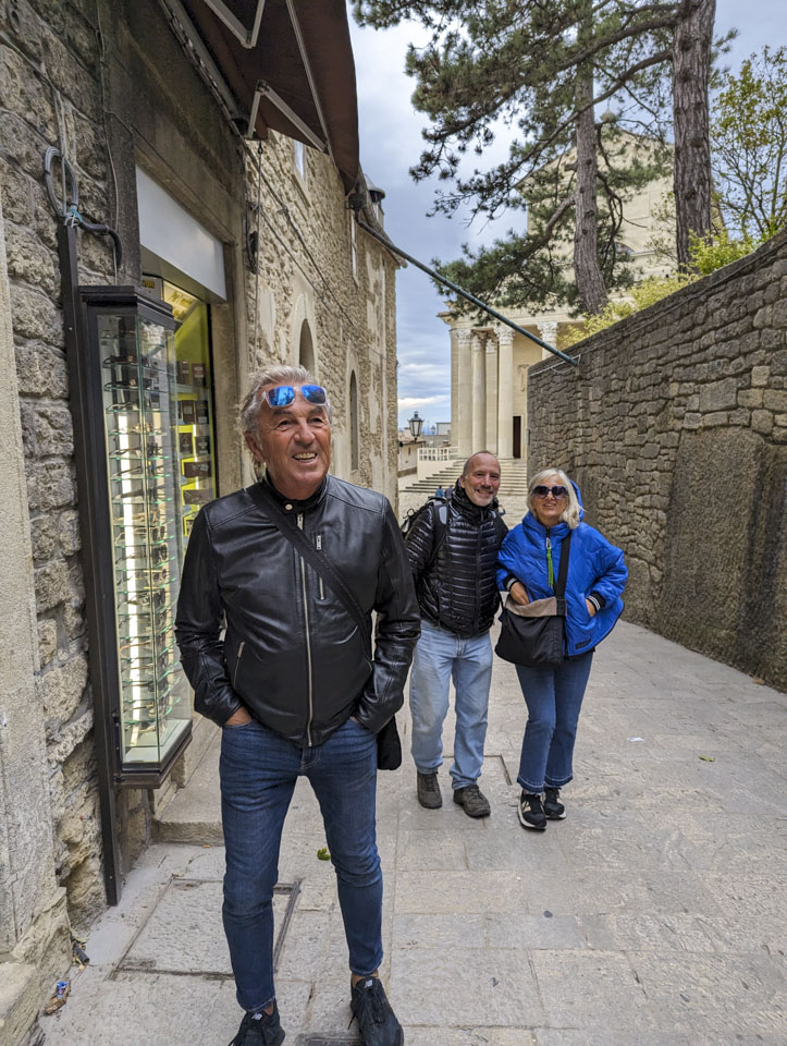 Roberto, Paul, and Alizia are in a narrow street that leads to the Basilica. An optician shop can be seen on the left, and a plain wall is on the right. 