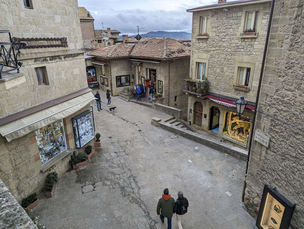 Looking down at a piazza. Two people are walking in from the bottom of the picture, and two other people are in the distance with a dog on a leash. There are stone buildings with shop windows. Alizia, Roberto, and Paul are just outside one of the shops.