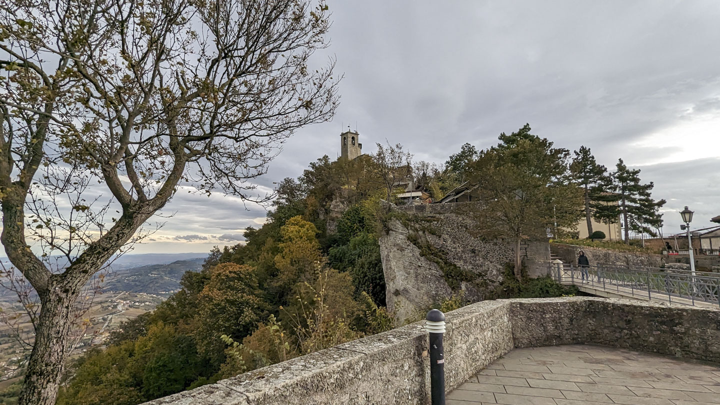 A tower can be seen at the top of a rise in the land. There is a walkway leading to it on the right. On the left is a tree, and far below is some land.