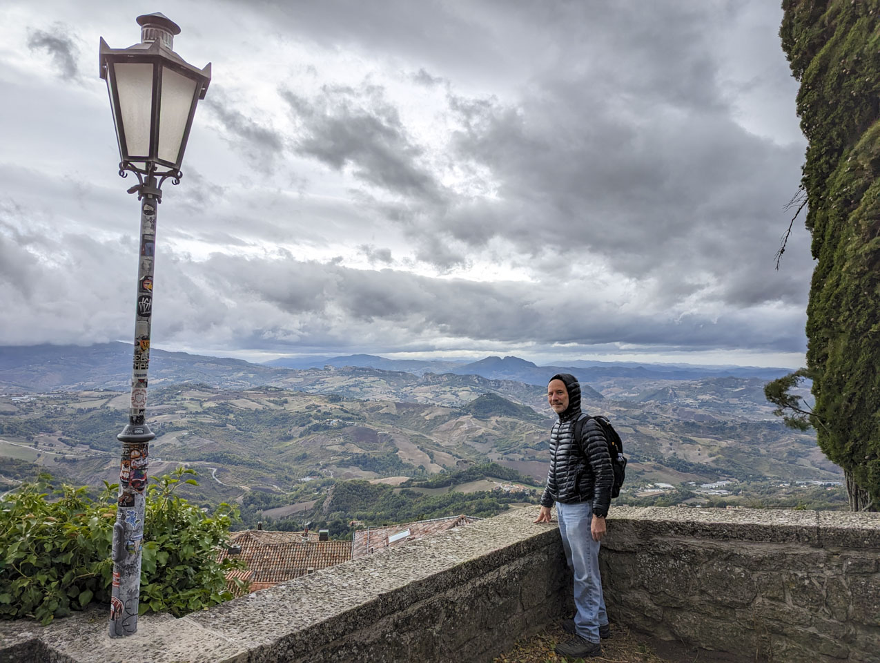 Paul is standing at the corner of a low stone wall. He is wearing a jacket with the hood up. The land on the other side of the wall drops off quickly.