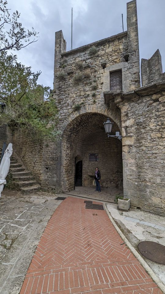 Anne is standing in a tall alcove in a stone wall, with a brick path that leads to the alcove. 