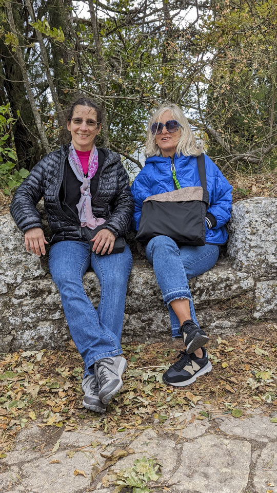 Anne and Alizia sit on a stone bench that is part of a low wall. The wind is blowing their hair.