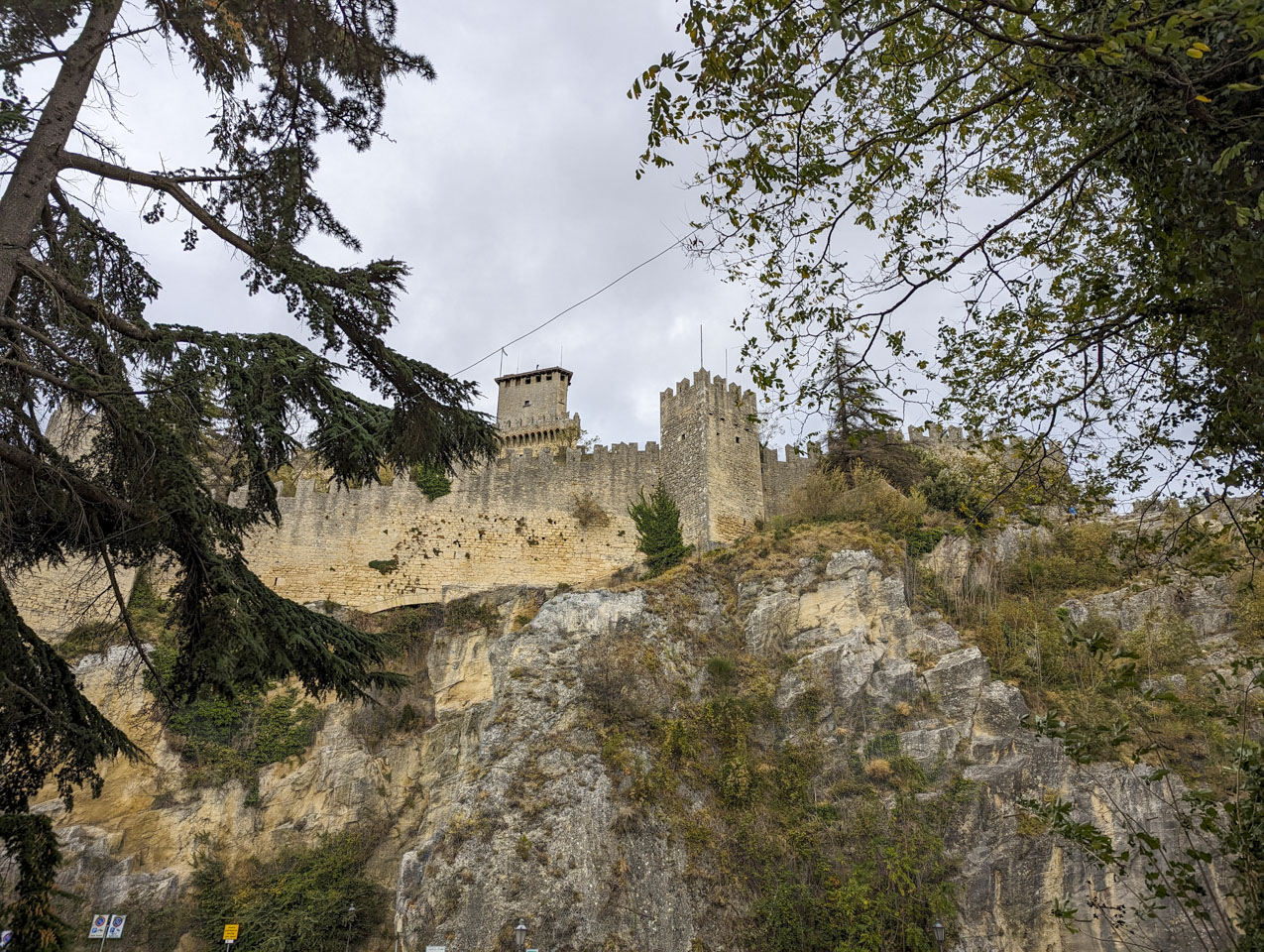 A tall rock cliff face with stone walls above it and the 1st tower seen even higher up.