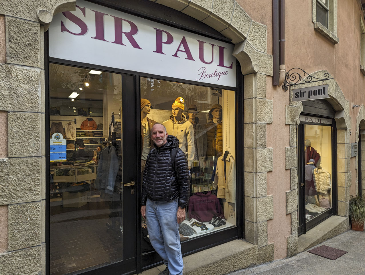 Paul stands in front of a store window with a sign above that says Sir Paul. Inside are men's clothing.