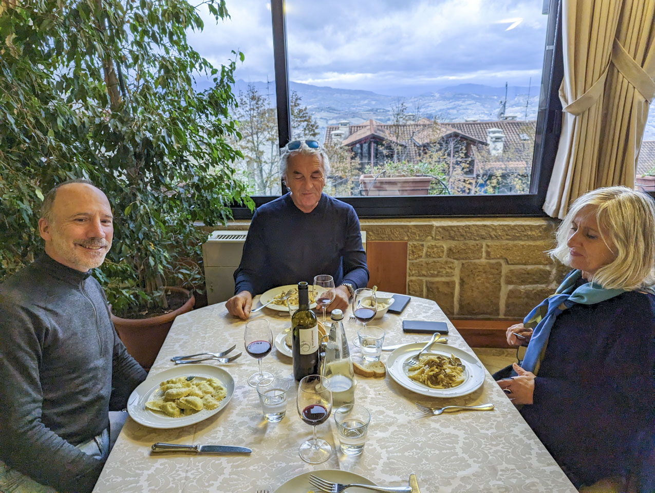 Paul, Robert, and Alizia at a table with a gold tablecloth. A plate of foot is in front of each of them. Through the window mountains can be seen in the distance.