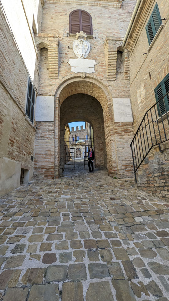 Looking up a steep stone walkway through a very large but narrow arch in a city wall. Anne is standing in front of an open grating.