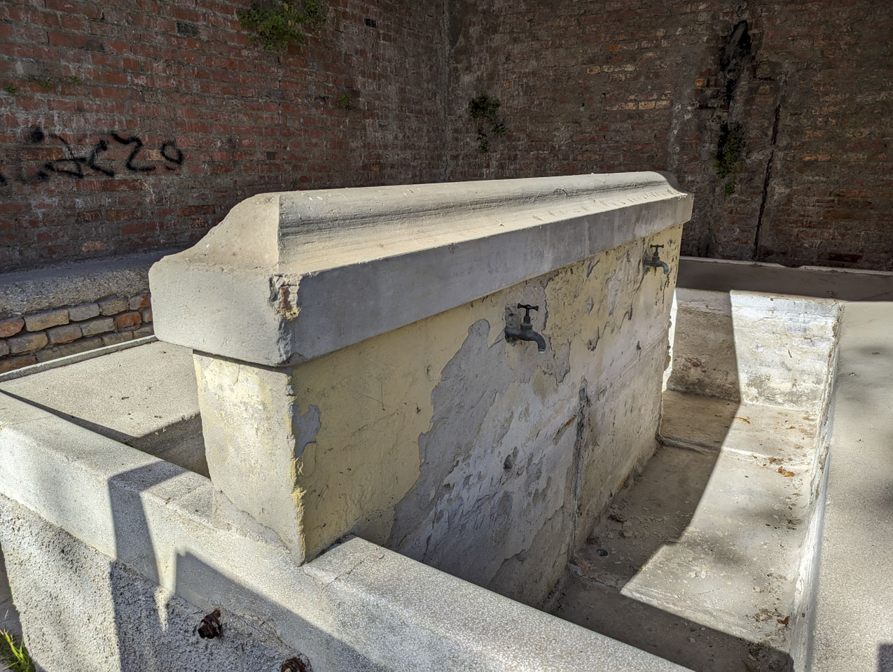 A cement barrier with water spouts that go into a trough area, inside a brick outdoor room.