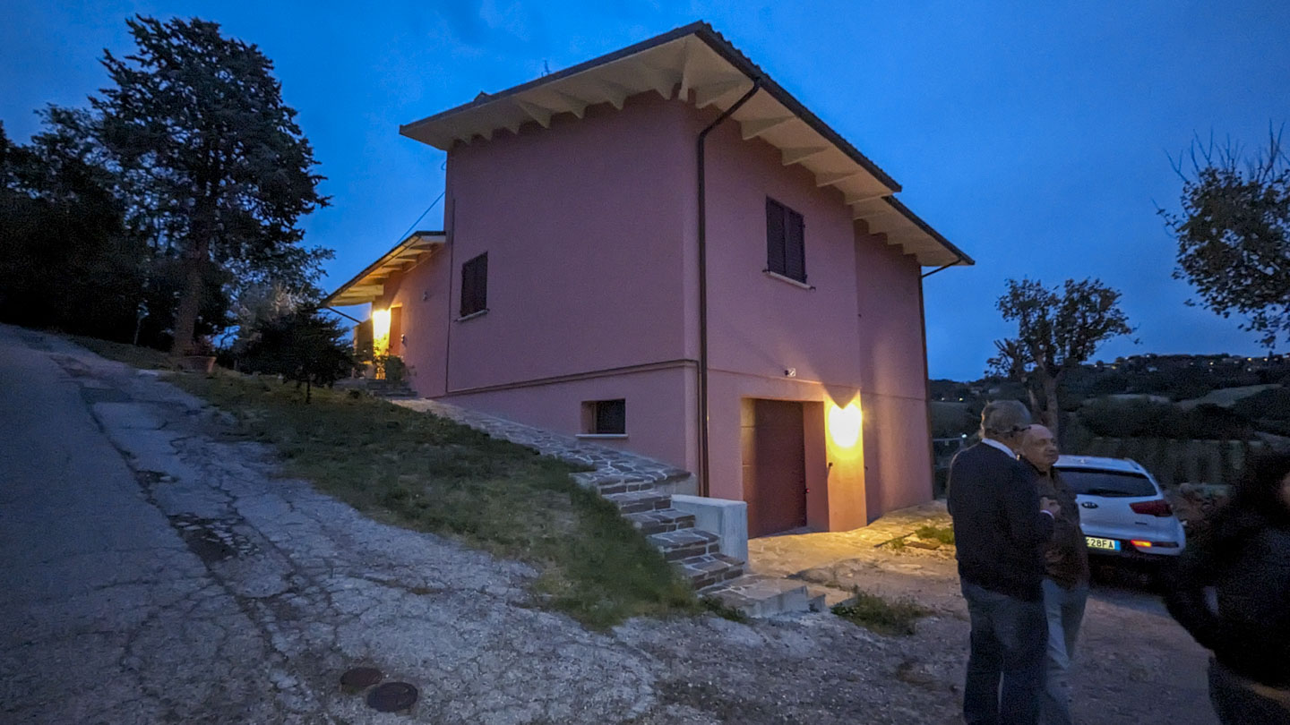 A clay colored house on a steep hill, with farm land and village lights in the background.