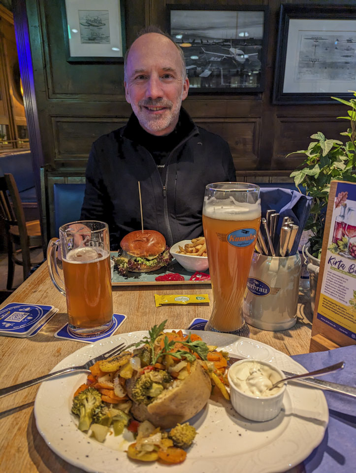 Paul sits at the far side of the table. In front of him is a rectangular plate with a burger and fries. Closer to us is a potato with broccoli, carrot, and other vegetables.