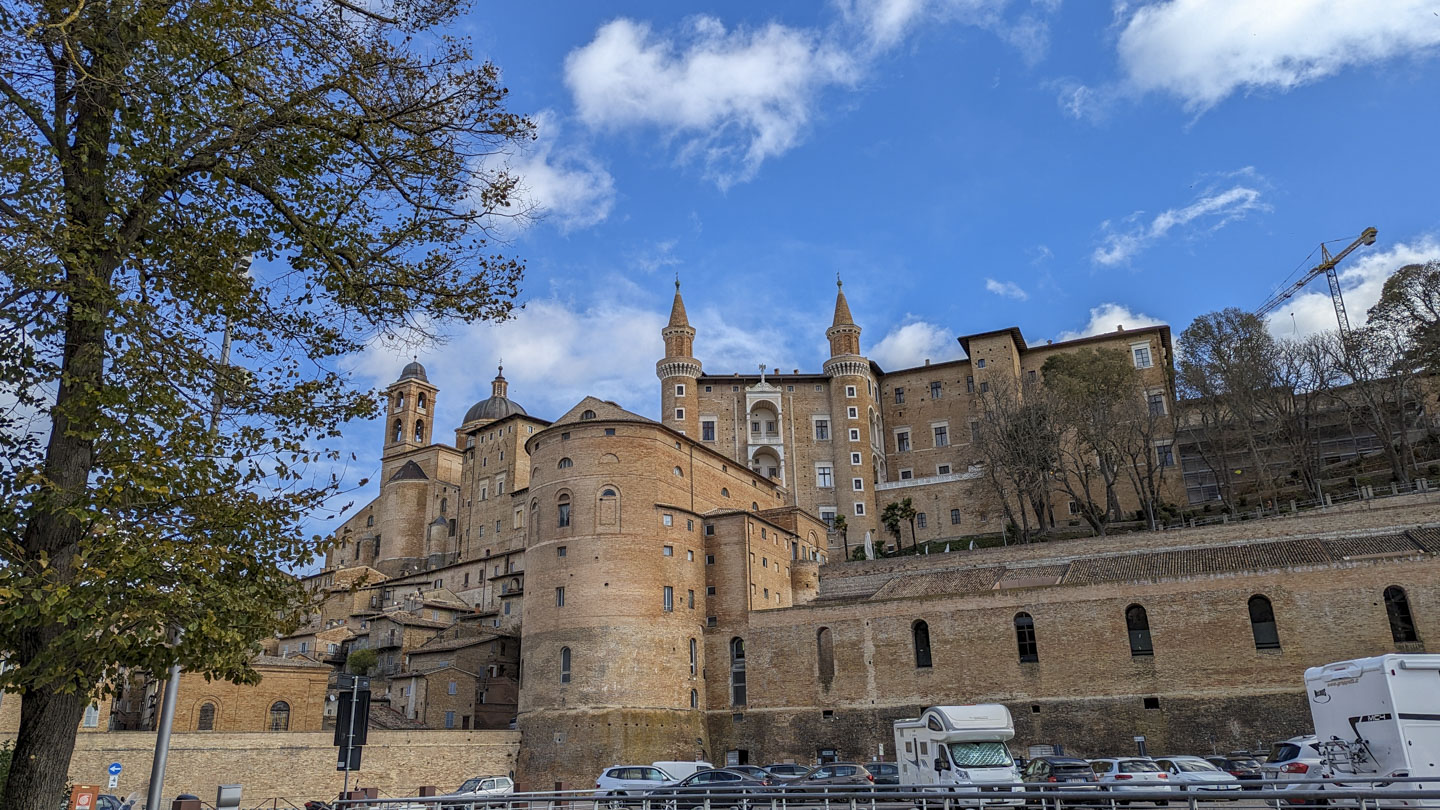 A sunny day with a few clouds, we can see a tree on the left. Behind some tall walls is a higher level of the city, where a majestically tall stone building stands.