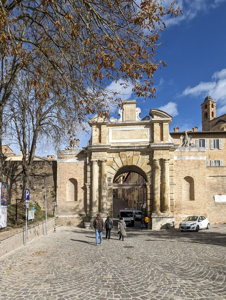 Three people, Francesco, Anne, and Doni, walk away from us towards an arched entrance in a tall stone wall.