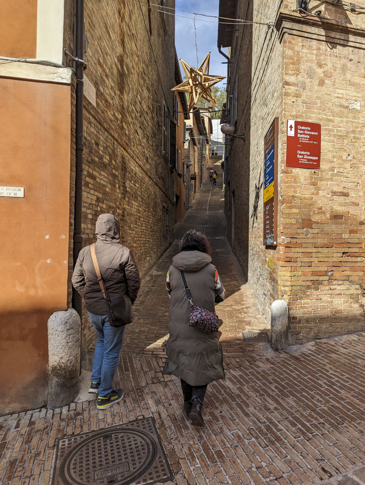 Francesco and Doni are entering a very narrow street between tall stone buildings. The street goes up, particularly steeply towards the far end.