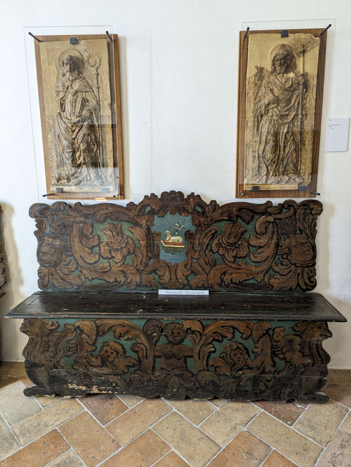 Two vertical reliefs of saints are on the wall, above a green bench with elaborate brown vines painted on it.