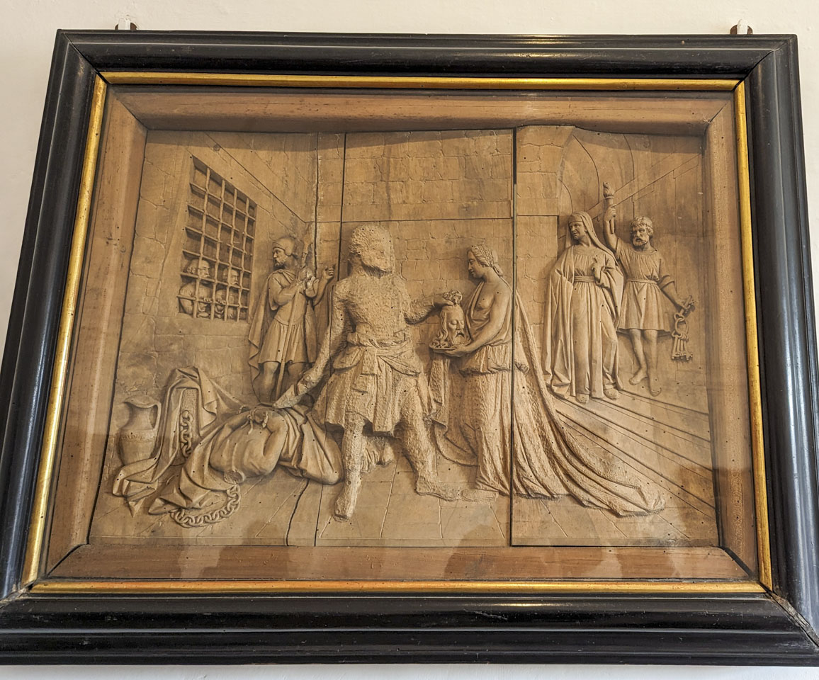 An intricately carve scene of a guard handing a decapitated head to a woman holding a tray. A couple of people look through a grated window and a second guard has turned to face them. On the other side is a robed man plus a jailor that is holding keys. The work is intricately done except the main guard and the dress of the woman.