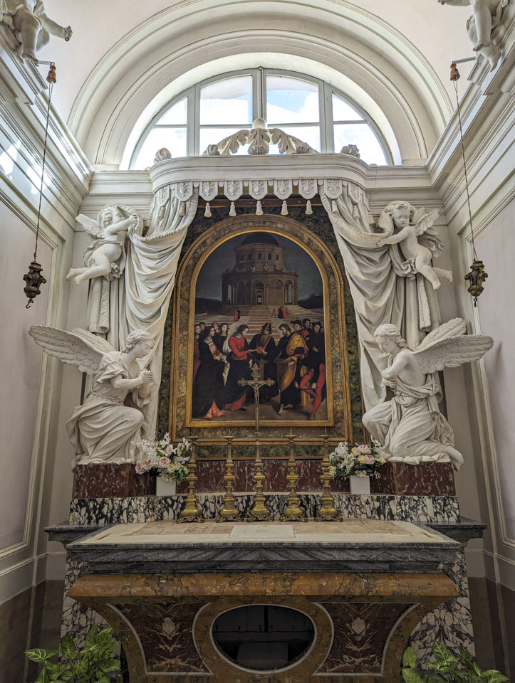 See description above. The sculpture has an angel kneeling on each side, and a cherub flying above them. There is an arched window in the wall above the sculpture.