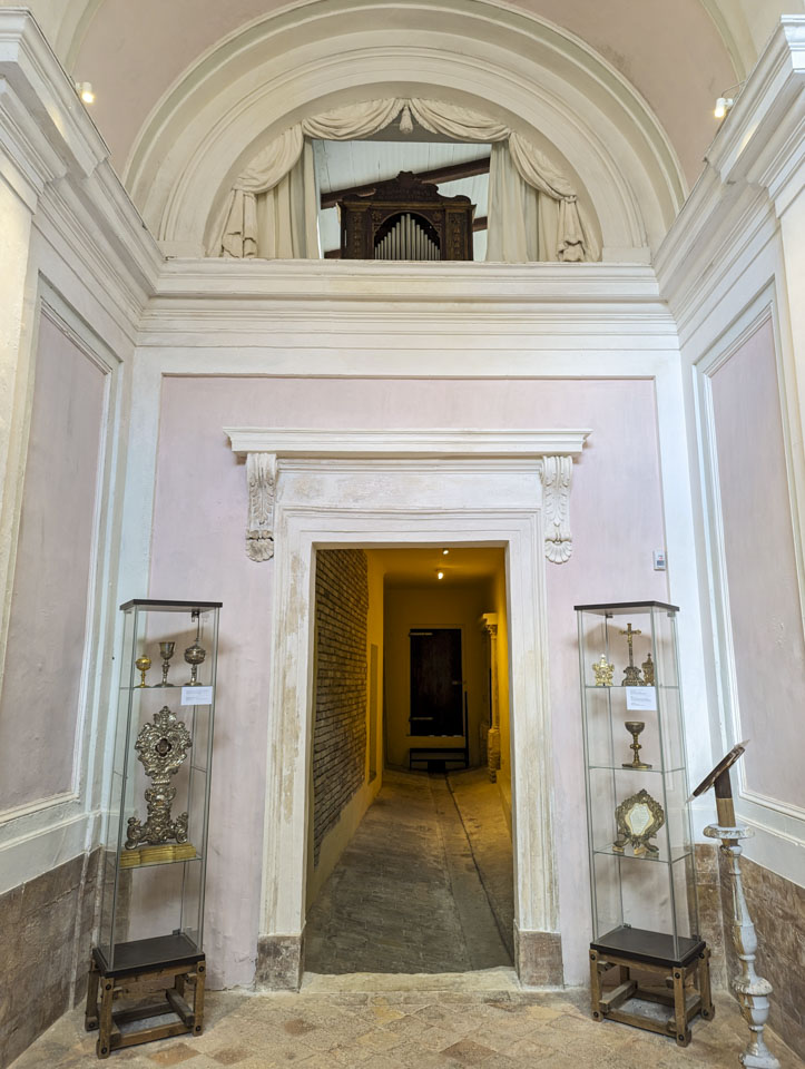 A room with pink walls and an open doorway. Through the doorway can be seen what once was an alleyway. It has a plain concrete floor and exposed brick.