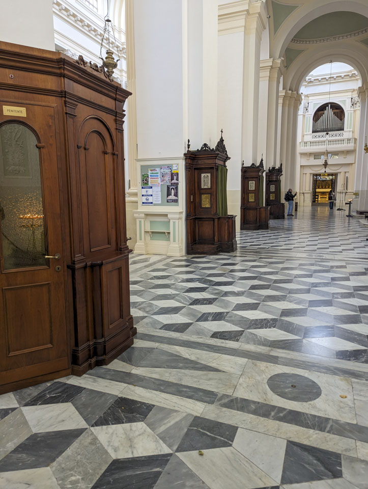 A wide aisle with four wooden confessionals against columns. Paul can be seen in the distance taking a photograph. Seen through the top of the archway are organ pipes.
