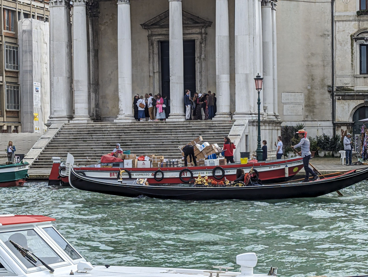 Looking over a canal in Venice to a building with many steps and people on the top of the stairs. In the water is a boat that is loaded with boxes, and there is a cart on the land next to it that is also loaded with boxes.