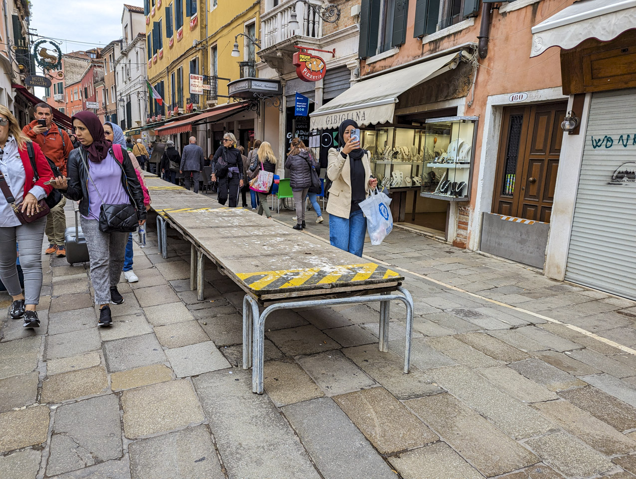 A street with shops on both sides, people walking, and in the center are wooden platforms available for when the road is flooded.