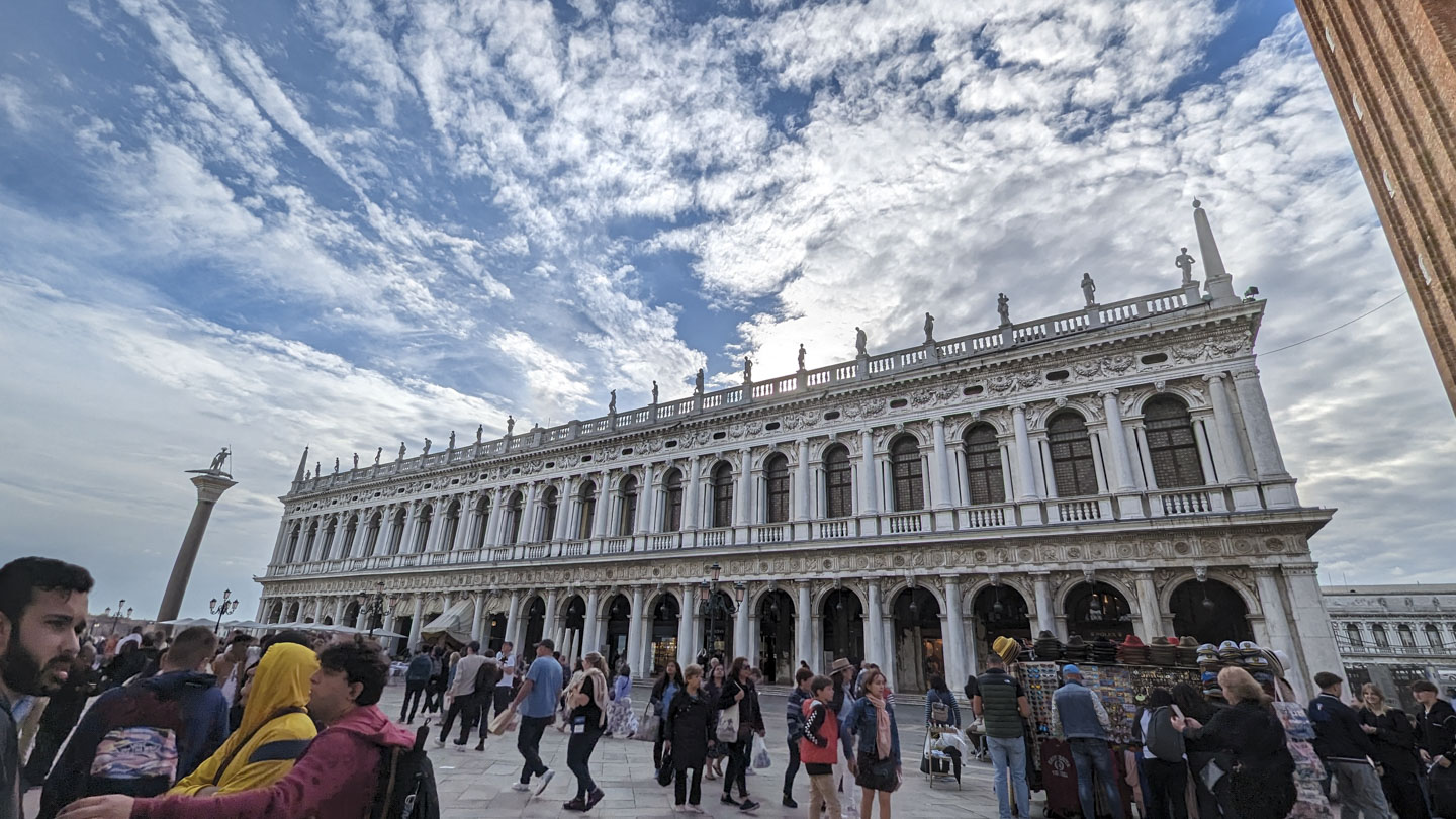 A blue sky with many white clouds is seen behind a two story very long building. The building has windows flanked by columns on the second floor, and archways flanked by columns on the first floor. There is a series of statues along the roofline.