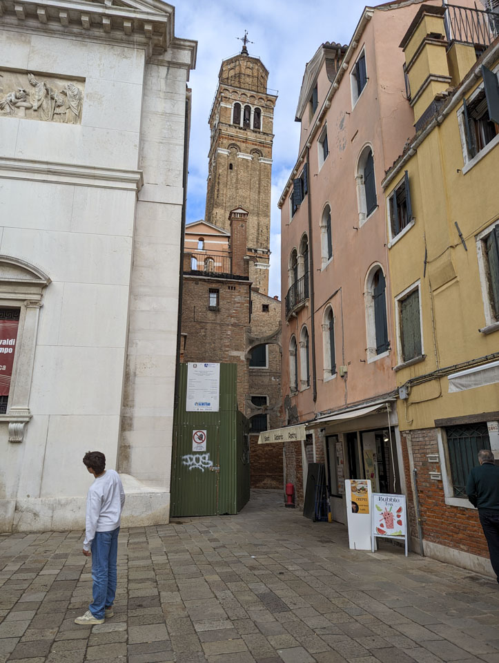 Looking from a small piazza towards a stone building with a tower. The lean of the tower is obvious because of the narrow area of visibility.
