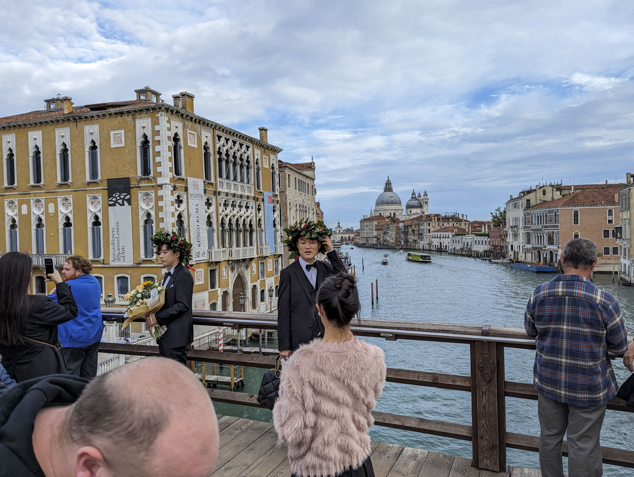 On a bridge in Venice, two men of oriental descent wear laurel leaf wreaths signifying that they graduated that day. Some friends take pictures of them.