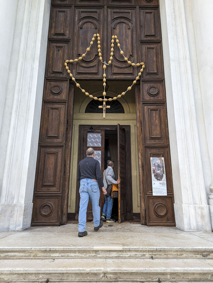 Very large entryway into a church. The dark wooden door has an oversized rosary hanging on it.