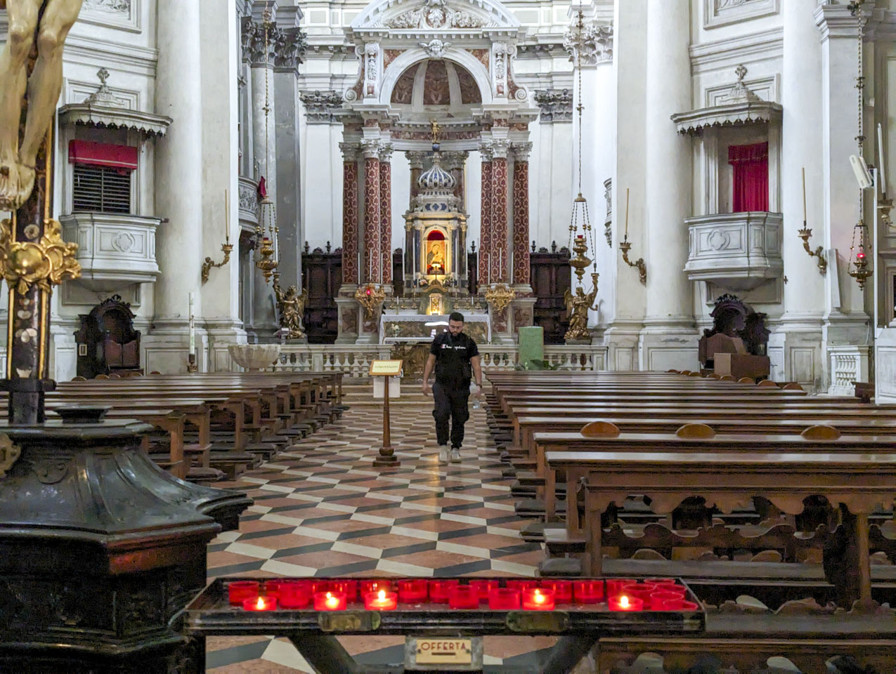 Interior of the church. Votive candles are in the foreground of the picture, then two columns of pews with a man walking down the aisle in the middle. The floor has different colored tiles that create the illusion of shallow raised platforms.