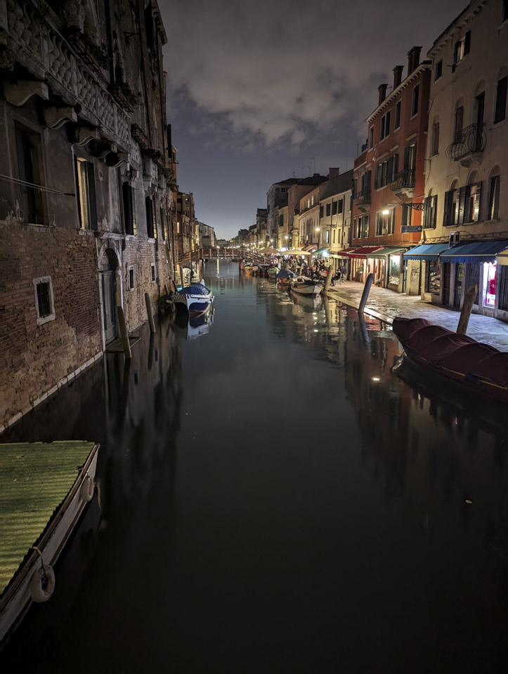 A night view down a canal in Venice. On the left the walls of the buildings go up to the water. On the right there is a walkway and many shops, including restaurants that are full of people.