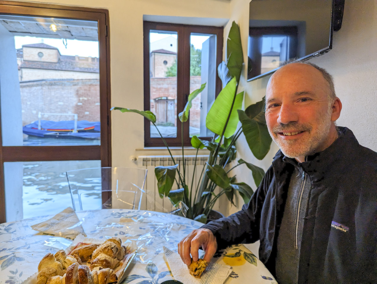 Paul sitting at a table that has some baked goods on it, with a Venetian canal visible through the window and glass door.