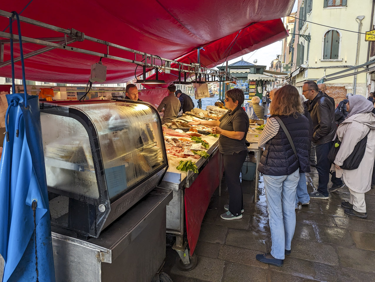 A red awning covers a fish stand full of ice and various seafood. Three men are behind the counter, on the left, while customers stand on the right.