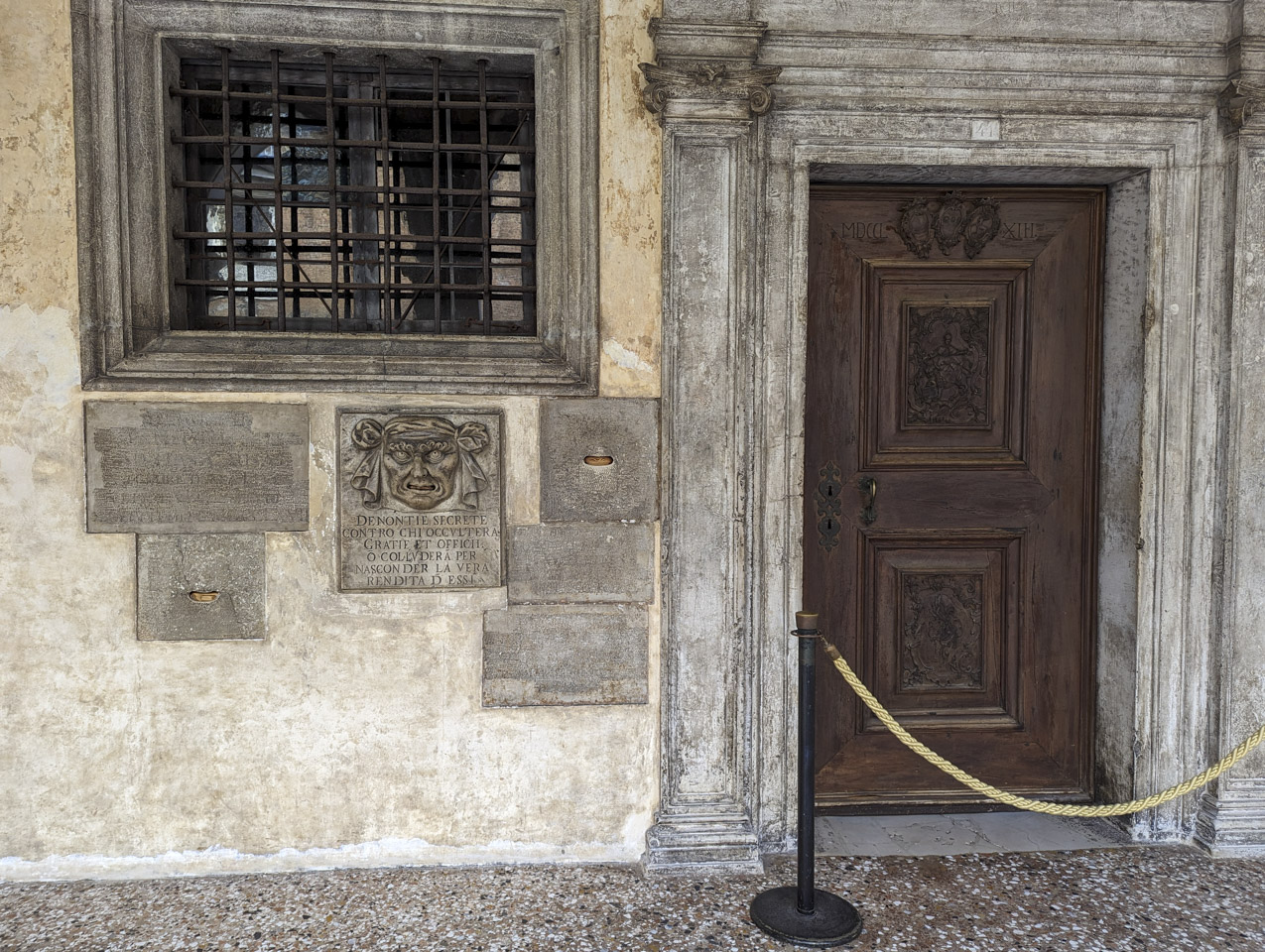 A wooden door is on the right, a window with iron lattice is in the upper left, and below it are many stone plaques plus one that still has a face carved on it.