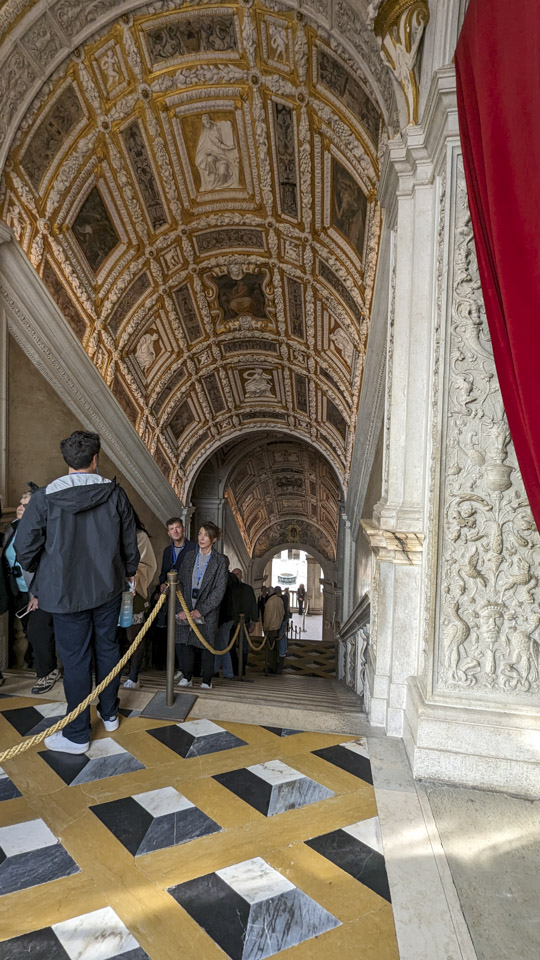 Looking down a large staircase, with many people going up on the left side. Some relief figures and other decorations can easily be seen on the ceiling of the stairwell.