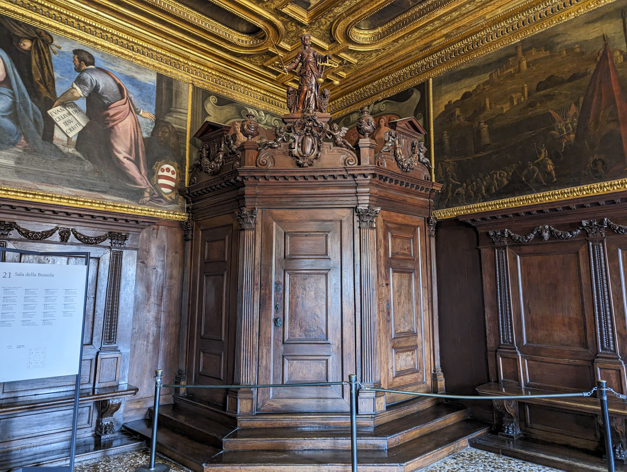 The corner of a room that has wood panels on the lower half of the walls and paintings above them. The ceiling is slightly less ornate than previous rooms but still gold colored. A wood structure with three sides fills the corner; at least 2 of the sides look like doors. A wooden statue of Justice with scales is on top of the structure.