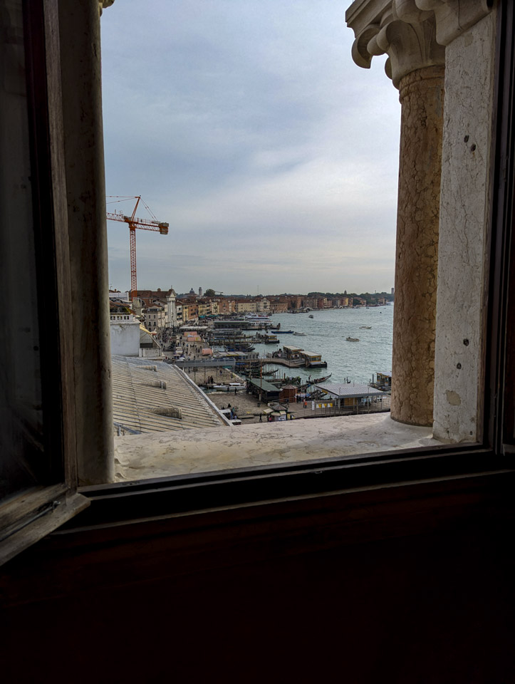Looking out a window in the Ducal Palace of Venice. Through the marble frame of the window can be seen buildings, water, boats, and some people.