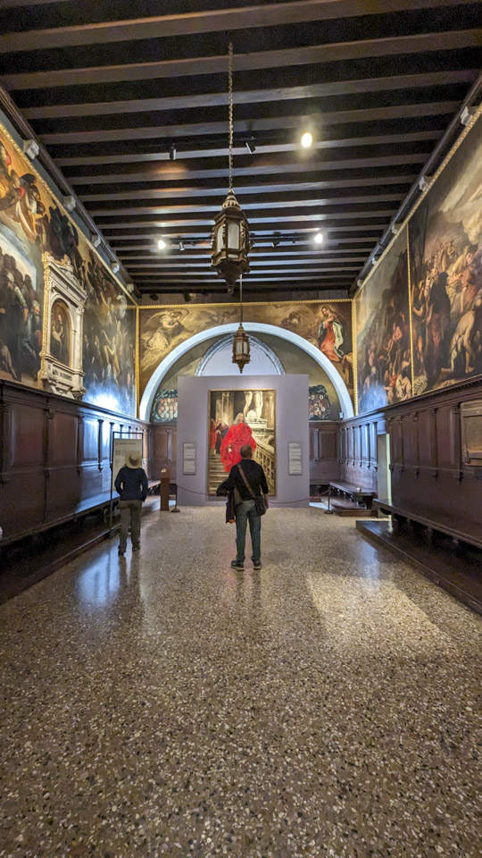Anne and Francesco standing in a large hallway with wood paneling and paintings.
