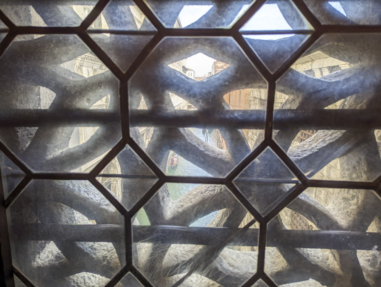 A window with glass through which is decorative stonework, and then buildings and a canal with a gondola can be seen.