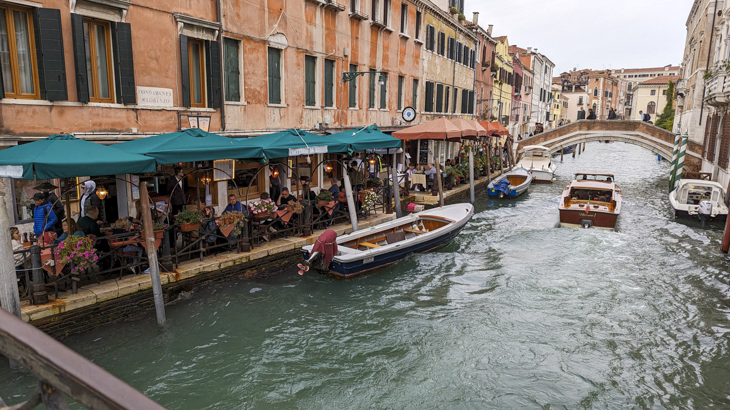A canal with tables along the side, some with green awnings and some with orange awnings. Every table has customers sitting at it.